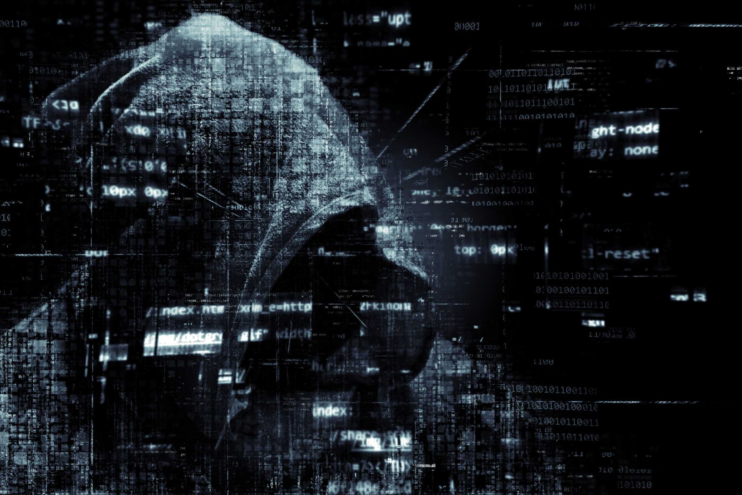 Unidentifiable person in a dark hoodie in front of a dark background, white writing and lines of code are recognizable in the foreground. The picture looks fragmented.